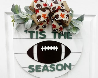 May include: A round wooden door hanger with a white background and green lettering that reads "'Tis the Season". A black football is in the center of the sign. A brown and white ribbon with football and leaf designs is tied above the sign.