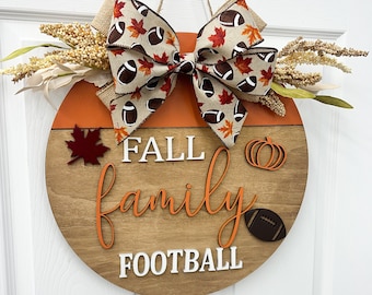 May include: A round wooden door hanger with the words "Fall family football" in orange and white lettering. The hanger is decorated with a brown and white ribbon with football and leaf designs, a brown and orange pumpkin, and a red maple leaf.