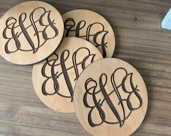 Może przedstawiać: Cztery okrągłe drewniane podkładki z wzorem monogramu. Podkładki są jasnobrązowe z ciemnobrązowym monogramem.