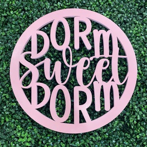 May include: A pink round wooden sign with the text "Dorm Sweet Dorm" cut out in the center.