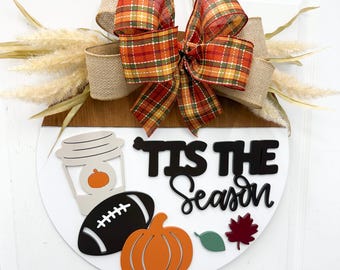 May include: A round wooden sign with a white and brown design, featuring a plaid ribbon bow and decorative elements. The sign includes a coffee cup, football, pumpkin, leaf, and the text "Tis the Season."