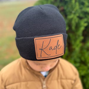 May include: Black knit beanie with a brown leather patch. The patch features the name "Kade" in a stylish script font. The beanie is a classic style, suitable for cold weather.