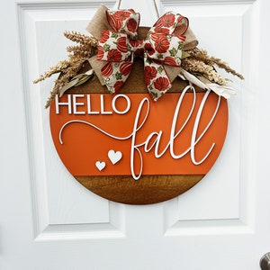 May include: A round wooden door sign featuring a dark brown bottom half and a bright orange top half. The white text reads HELLO fall with two small white hearts. A burlap and pumpkin-patterned ribbon bow sits at the top with dried wheat stalks.
