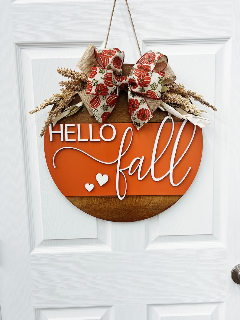 May include: A round wooden door sign featuring a dark brown bottom half and a bright orange top half. The white text reads HELLO fall with two small white hearts. A burlap and pumpkin-patterned ribbon bow sits at the top with dried wheat stalks.