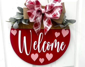 May include: A red and white checkered bow with burlap accents hangs from a round wooden door hanger. The door hanger is painted red and has the word "Welcome" in white lettering. There are pink hearts scattered around the word.