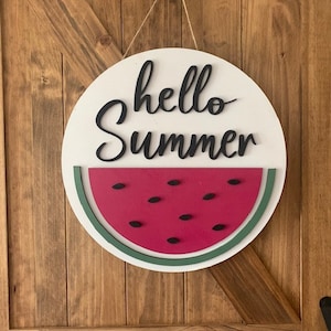 以下が含まれることがあります： 白木の丸い看板に、黒い文字で「hello summer」と書いてあり、緑とピンクのスイカのグラフィックが描かれています。