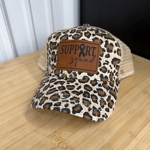 May include: A leopard print trucker hat with a brown leather patch that reads "SUPPORT squad" with a black ribbon graphic. The hat has a tan mesh back and a curved brim.