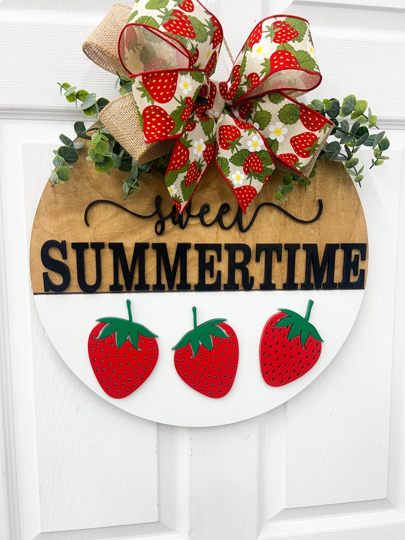 May include: Round wooden sign with the words "sweet SUMMERTIME" in black. The sign features three red strawberries with green stems and a decorative bow with a strawberry pattern.