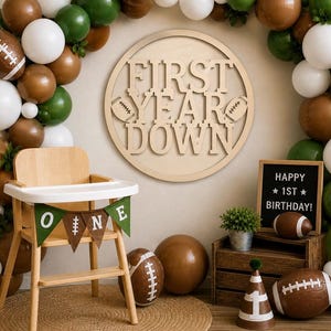 May include: A wooden high chair with a banner that says "ONE" is set up for a first birthday party. The party decorations include a balloon arch in brown, white, and green, a wooden sign that says "FIRST YEAR DOWN", and football-themed decorations.