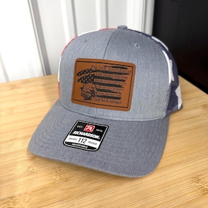 May include: A gray trucker hat with a brown leather patch featuring the text "The 2nd Amendment" and an eagle graphic. The hat has a mesh back and a curved brim. The Richardson brand label is visible.