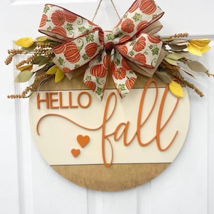 May include: A round wooden door sign featuring the text HELLO fall in orange 3D lettering with two small orange hearts. The sign is decorated with a large burlap bow patterned with orange pumpkins and green leaves, accented by yellow and brown foliage.
