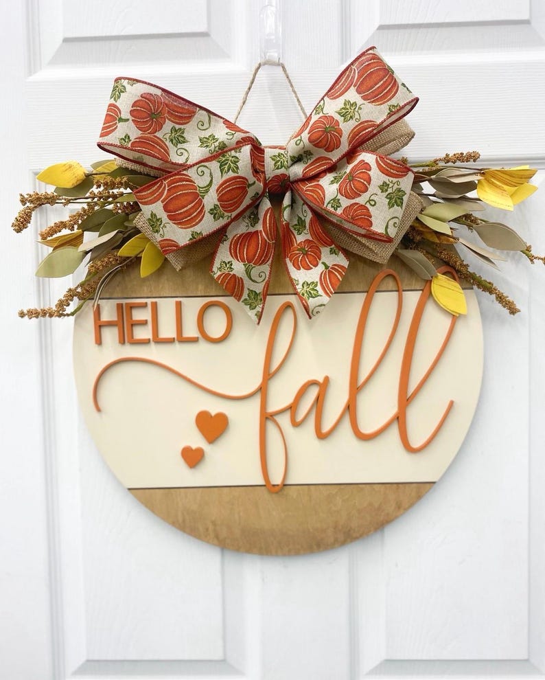 May include: A round wooden door sign featuring the text HELLO fall in orange 3D lettering with two small orange hearts. The sign is decorated with a large burlap bow patterned with orange pumpkins and green leaves, accented by yellow and brown foliage.