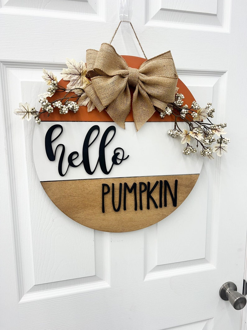 May include: A round wooden door hanger with a white and brown wood grain design. The top half is orange and the bottom half is white. The words "hello pumpkin" are written in black on the door hanger. The door hanger is decorated with a burlap bow and white berries.