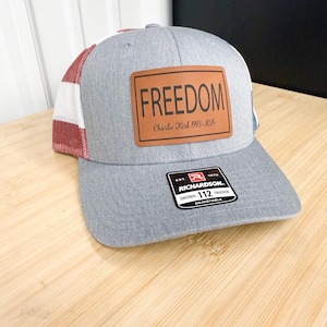 May include: Gray trucker hat with a brown leather patch that reads "FREEDOM" and "Charlie Kirk 1993-2015." The hat has a red and white striped mesh back and a black and white tag that reads "RICHARDSON ORIGINAL 112 TRUCKER ADJUSTABLE."