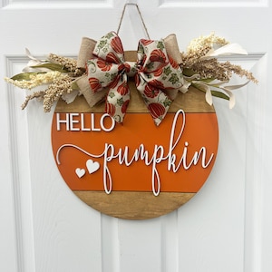 May include: A round wooden door hanger with a brown and orange striped design. The hanger has the words "Hello pumpkin" written in white cursive lettering with two white hearts. A burlap bow with a pumpkin print ribbon is attached to the top of the hanger.