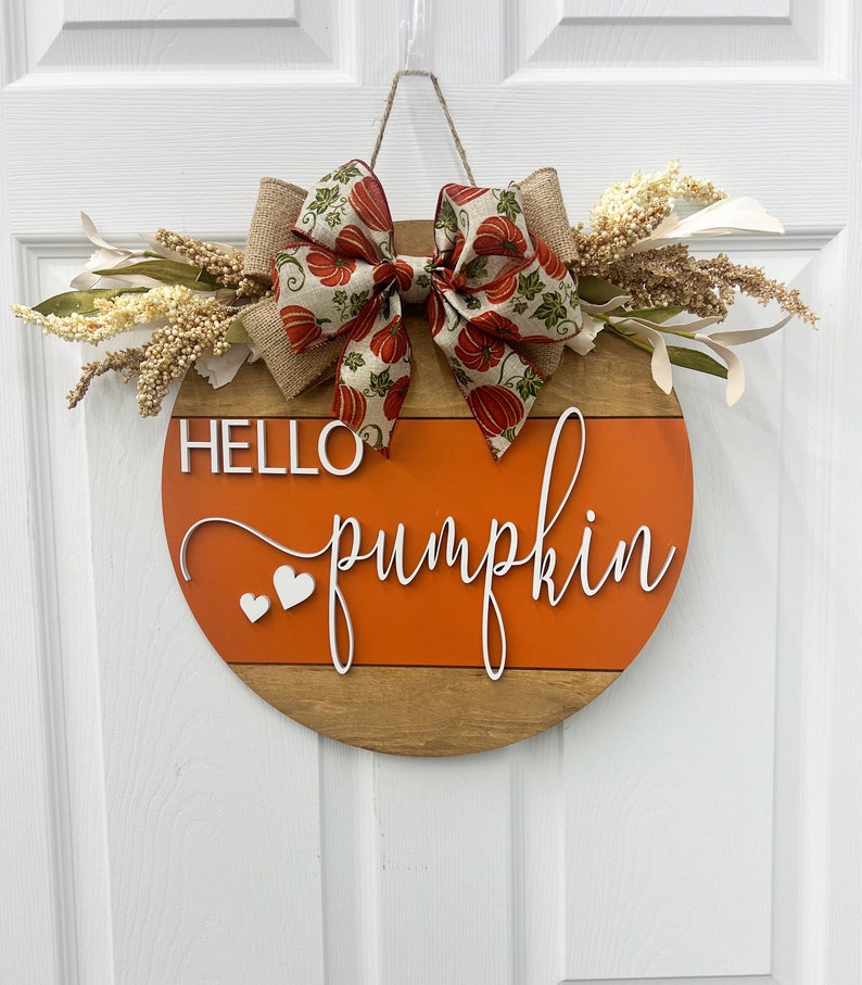 May include: A round wooden door hanger with a brown and orange striped design. The hanger has the words "Hello pumpkin" written in white cursive lettering with two white hearts. A burlap bow with a pumpkin print ribbon is attached to the top of the hanger.
