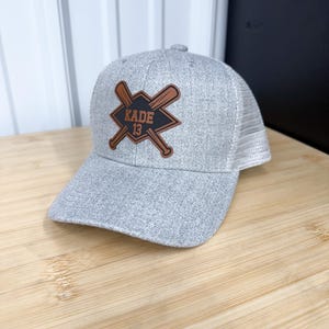 May include: Gray baseball cap with a brown leather patch featuring crossed baseball bats and the text "KADE 13".