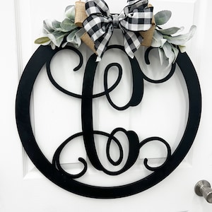 May include: Black and white checkered bow with burlap accents and greenery on a black wooden wreath with a large black metal letter "L" in a decorative script font.