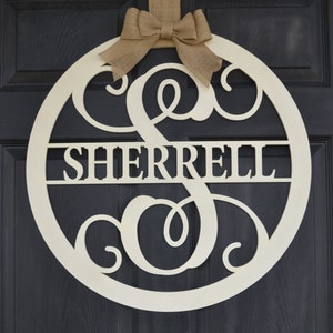May include: A white wooden round door hanger with the name "SHERRELL" and a large "S" initial in a decorative script font. The hanger is decorated with a burlap bow and is hanging on a black door.