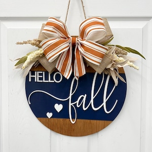 May include: A round wooden door sign featuring a dark blue and natural wood finish. The sign displays the white text HELLO fall and two small white hearts. It is decorated with a large orange and white striped bow and dried floral accents.