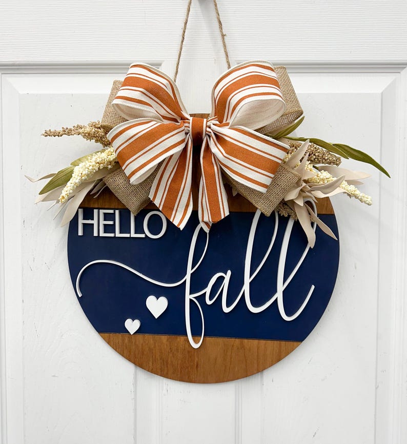 May include: A round wooden door sign featuring a dark blue and natural wood finish. The sign displays the white text HELLO fall and two small white hearts. It is decorated with a large orange and white striped bow and dried floral accents.