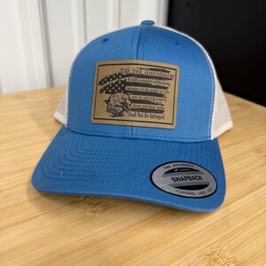 May include: Blue and white trucker hat with a curved brim. The hat features a rectangular patch with text about the 2nd Amendment and an eagle graphic. The hat has a snapback closure.