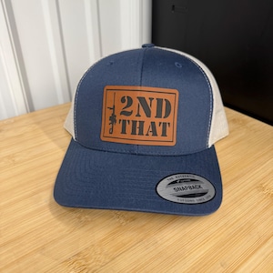 May include: Navy blue and tan trucker hat with a brown leather patch. The patch features the text "2ND THAT" with a rifle graphic. The hat has a curved brim and a snapback closure. The tag reads "The Authentic Snapback".