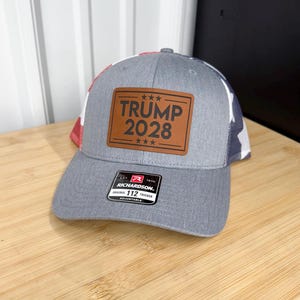 May include: Gray trucker hat with a brown leather patch that reads "TRUMP 2028" in white lettering. The hat has a white mesh back and a red and blue patterned side. The Richardson brand label is visible.
