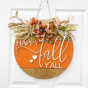 May include: A round wooden door hanger with an orange background and the text "Happy fall y'all" in white. The hanger is decorated with a burlap bow with a fall leaf print and a sprig of dried flowers.