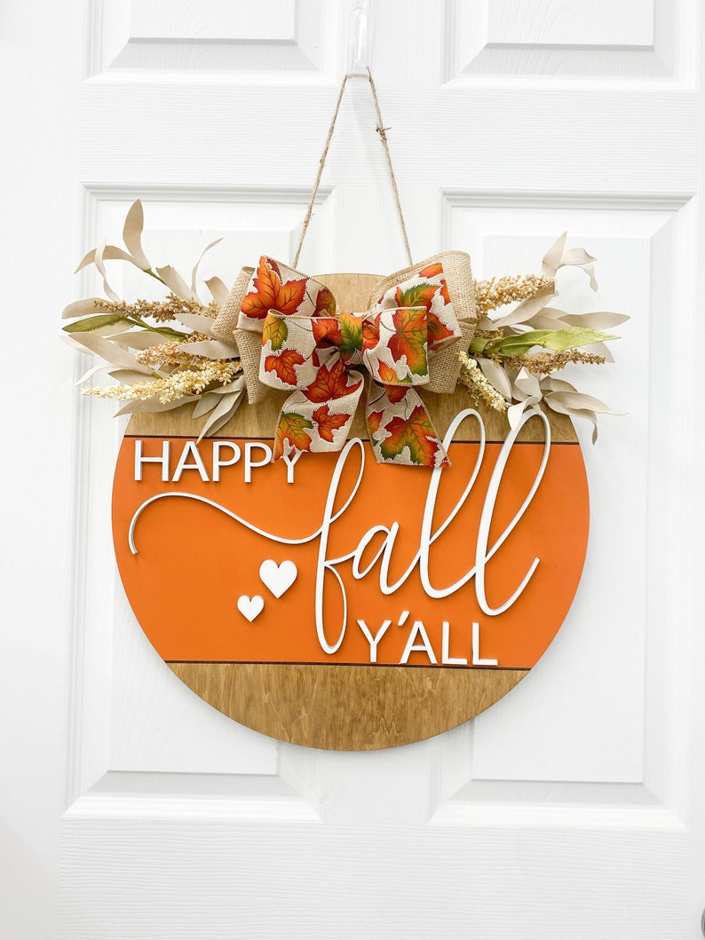 May include: A round wooden door hanger with an orange background and the text "Happy fall y'all" in white. The hanger is decorated with a burlap bow with a fall leaf print and a sprig of dried flowers.