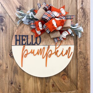 May include: A round wooden sign with the words "Hello Pumpkin" in orange lettering. The sign is decorated with a large bow made of orange, plaid, and burlap ribbon, and sprigs of greenery.