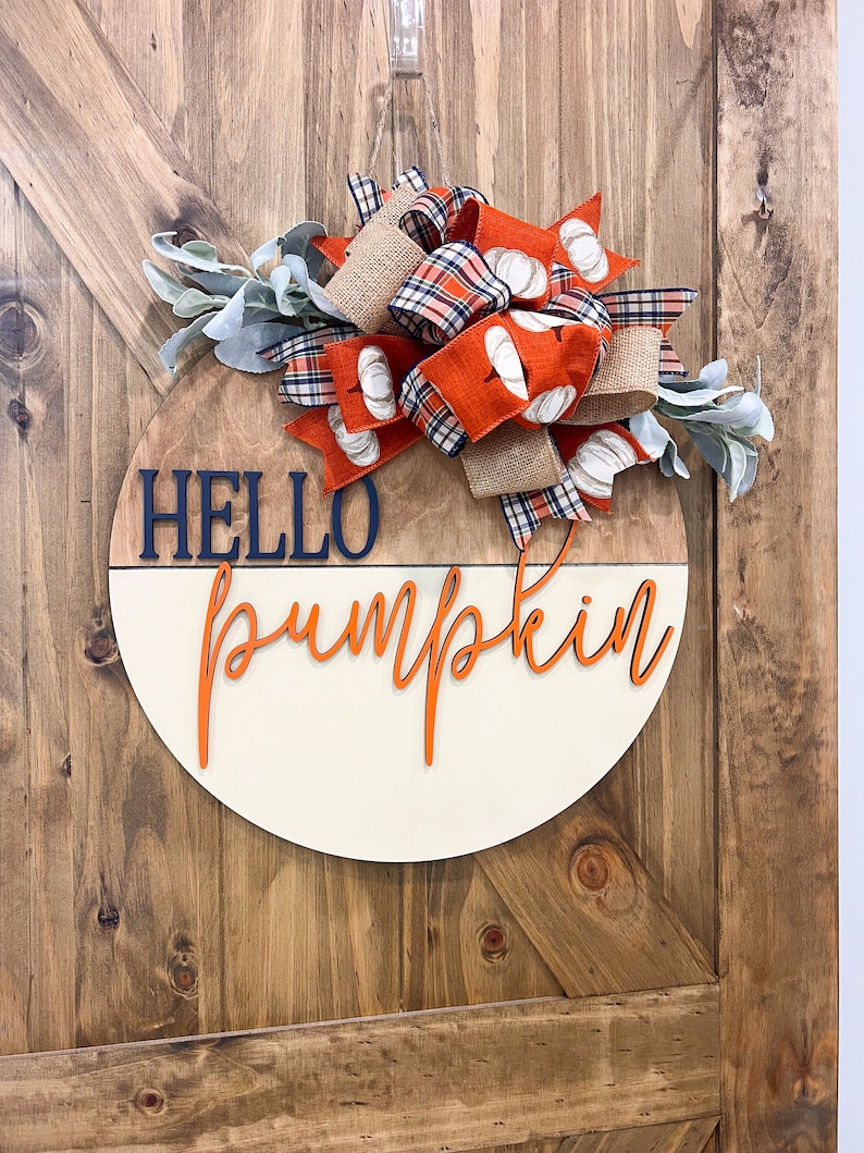 May include: A round wooden sign with the words "Hello Pumpkin" in orange lettering. The sign is decorated with a large bow made of orange, plaid, and burlap ribbon, and sprigs of greenery.