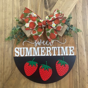 May include: Round wooden door hanger with a strawberry theme. The top half is wood-toned, the bottom half is black. It features the words "sweet summertime" and three red strawberries. A bow with a strawberry pattern and greenery complete the design.