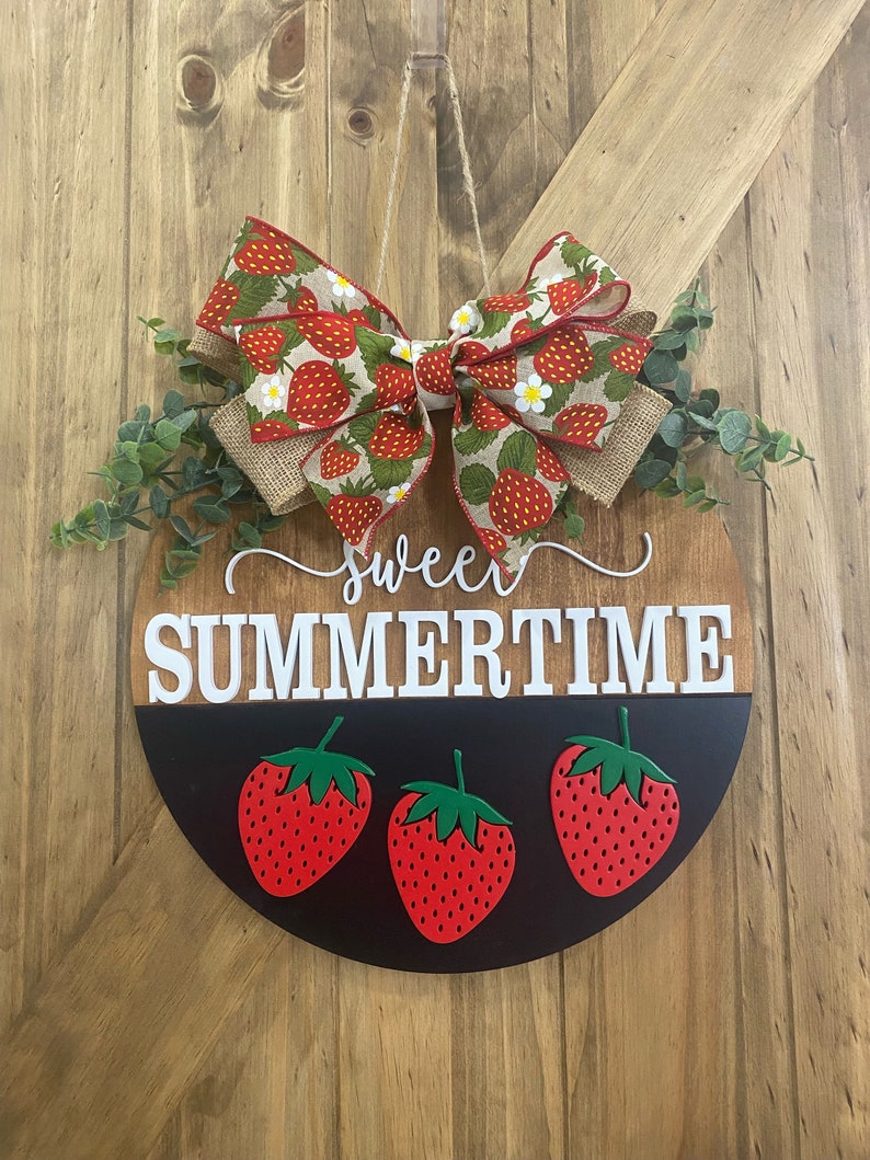 May include: Round wooden door hanger with a strawberry theme. The top half is wood-toned, the bottom half is black. It features the words "sweet summertime" and three red strawberries. A bow with a strawberry pattern and greenery complete the design.