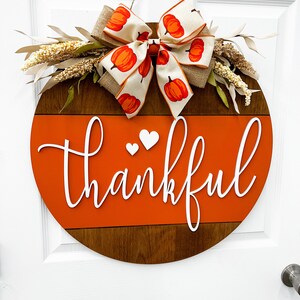 May include: A round wooden door hanger with a brown and orange painted design. The top half of the door hanger is brown wood with a white painted stripe. The bottom half is orange with the word "thankful" written in white. A white bow with a pumpkin pattern is attached to the top of the door hanger.