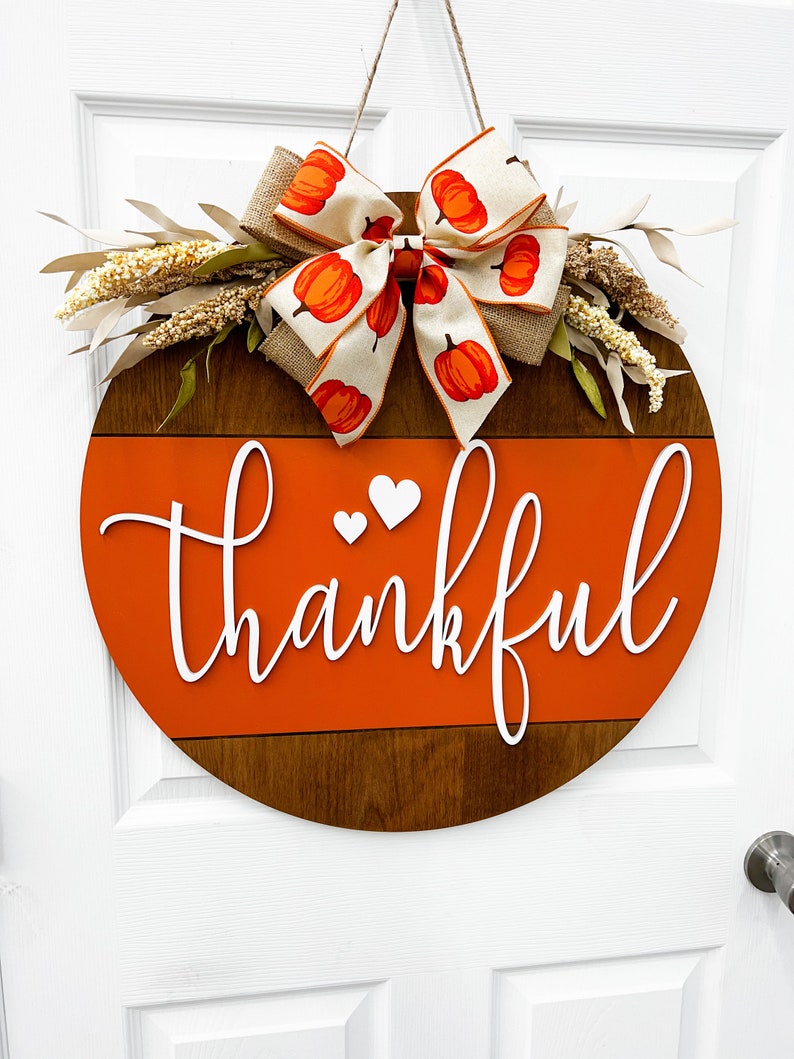 May include: A round wooden door hanger with a brown and orange painted design. The top half of the door hanger is brown wood with a white painted stripe. The bottom half is orange with the word "thankful" written in white. A white bow with a pumpkin pattern is attached to the top of the door hanger.