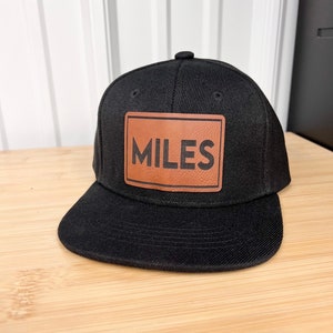 以下が含まれることがあります： 「MILES」と書いてある茶色の革のパッチが付いた黒い野球帽です。