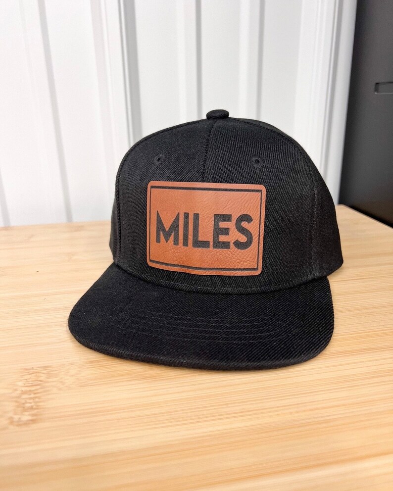 以下が含まれることがあります： 「MILES」と書いてある茶色の革のパッチが付いた黒い野球帽です。