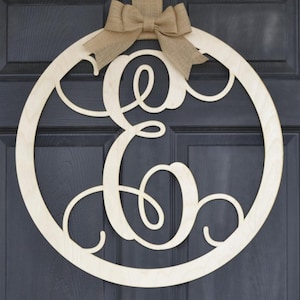 May include: A wooden door wreath with a large cursive letter "E" cut out in the center. The wreath is hung on a dark gray door with a burlap ribbon and bow.