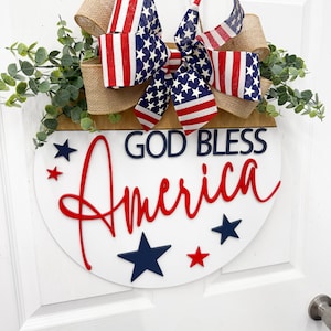 Puede incluir: Un letrero de puerta redondo de madera con el texto GOD BLESS en azul oscuro y America en letra roja. El letrero est&aacute; decorado con un lazo grande hecho de arpillera y cinta con patr&oacute;n de bandera estadounidense, junto con ramas de eucalipto verdes y estrellas azul marino y rojas.