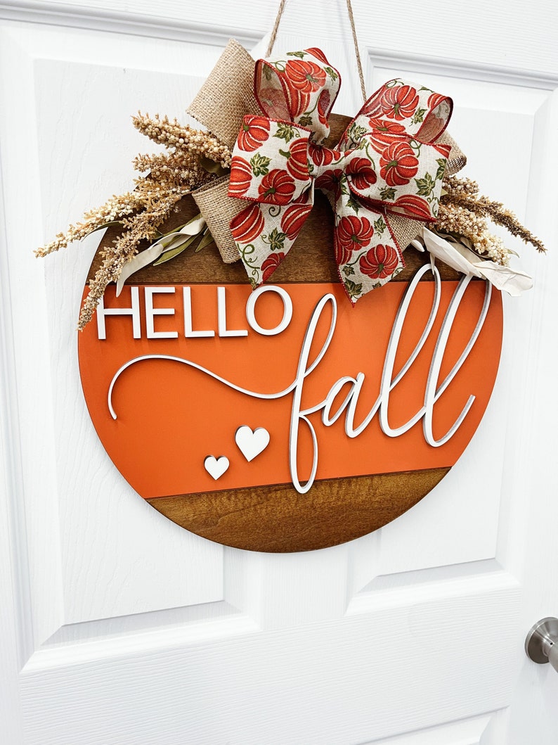 May include: A round wooden door sign featuring an orange horizontal panel with the white 3D text HELLO fall and two small white hearts. The sign is decorated with a large bow featuring a pumpkin pattern, burlap ribbon, and dried wheat stalks.