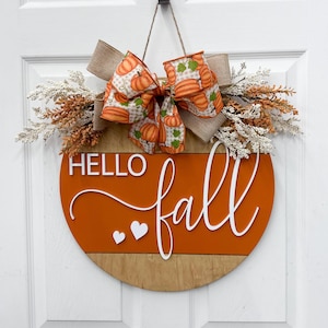 May include: A round wooden door sign featuring the text HELLO fall in white lettering with two small white hearts. The sign is decorated with a large bow patterned with orange pumpkins and tan burlap, accented by sprigs of white and orange foliage.