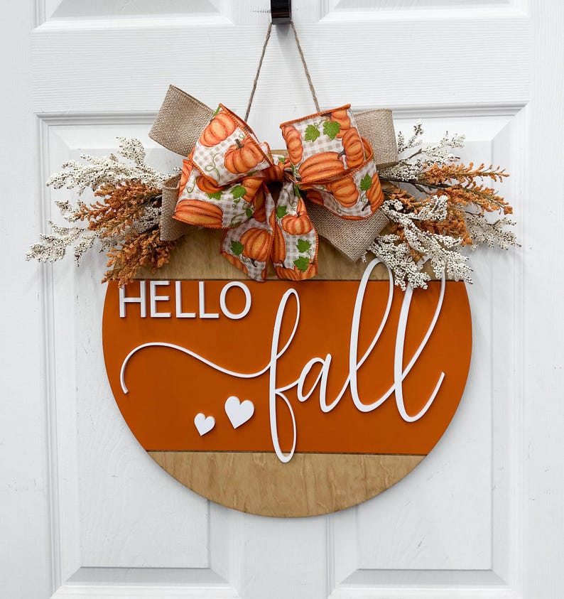 May include: A round wooden door sign featuring the text HELLO fall in white lettering with two small white hearts. The sign is decorated with a large bow patterned with orange pumpkins and tan burlap, accented by sprigs of white and orange foliage.