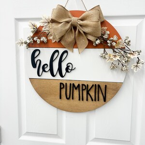 May include: A round wooden door hanger with a brown, white, and orange color scheme. The top half of the hanger is orange, the middle is white, and the bottom is brown. The words "hello pumpkin" are written in black on the white and brown sections. A burlap bow and white berry sprigs are attached to the top of the hanger.