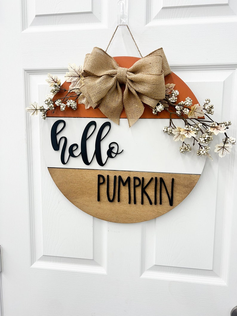 May include: A round wooden door hanger with a brown, white, and orange color scheme. The top half of the hanger is orange, the middle is white, and the bottom is brown. The words "hello pumpkin" are written in black on the white and brown sections. A burlap bow and white berry sprigs are attached to the top of the hanger.