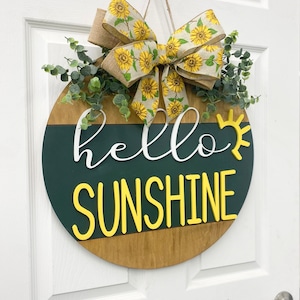 May include: A round wooden door hanger with a green and brown background. The words "hello sunshine" are written in white and yellow letters. A sunflower patterned bow and greenery are attached to the top.
