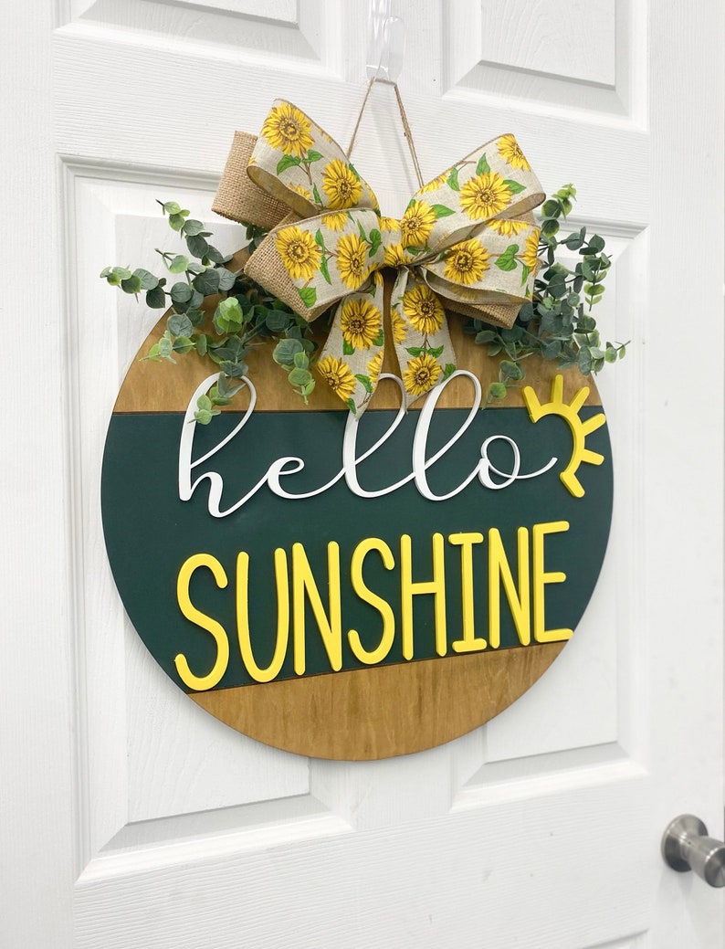 May include: A round wooden door hanger with a green and brown background. The words "hello sunshine" are written in white and yellow letters. A sunflower patterned bow and greenery are attached to the top.