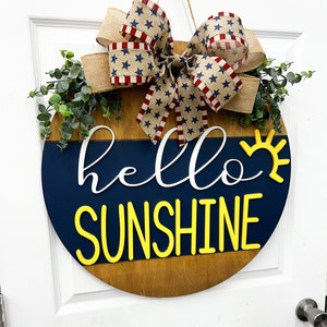 May include: A wooden round door hanger with a blue and brown background. The words "hello sunshine" are written in white and yellow letters. A red, white, and blue bow with stars is attached to the top of the door hanger.