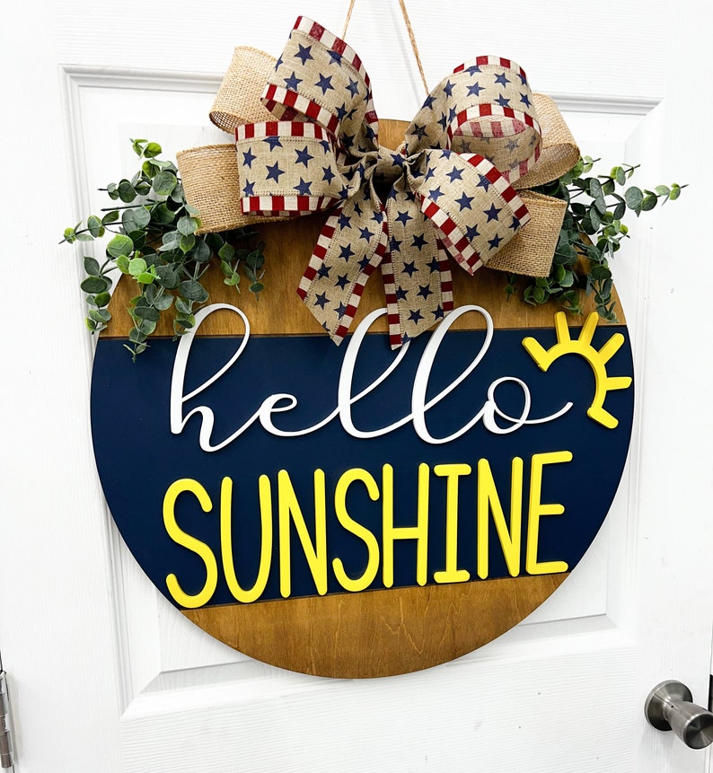 May include: A wooden round door hanger with a blue and brown background. The words "hello sunshine" are written in white and yellow letters. A red, white, and blue bow with stars is attached to the top of the door hanger.