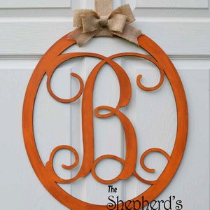 May include: A round, orange wooden door hanger with a large, decorative, cursive letter "B" in the center. The hanger is adorned with a burlap bow and has the text "The Shepherd's Wood Works & Engraving Hand" at the bottom.